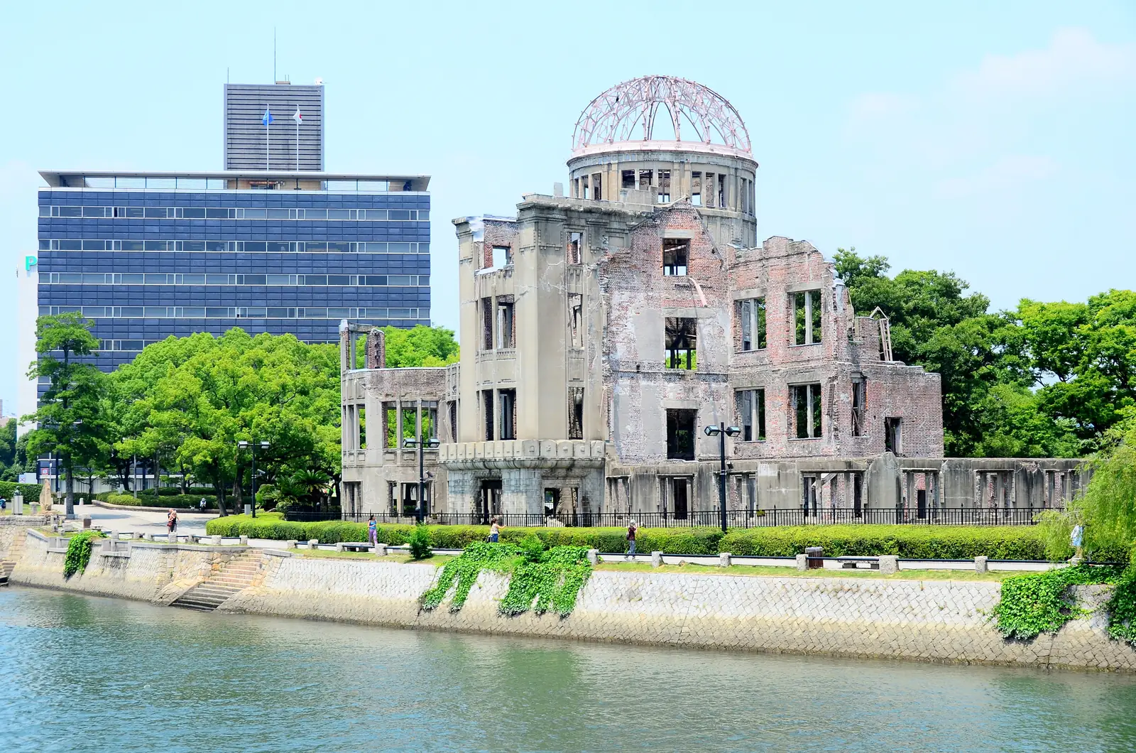 The ruined Atomic Bomb Dome with its bare steel dome frame, seen across the Motoyasu River on a sunny day, with green trees and an office block behind