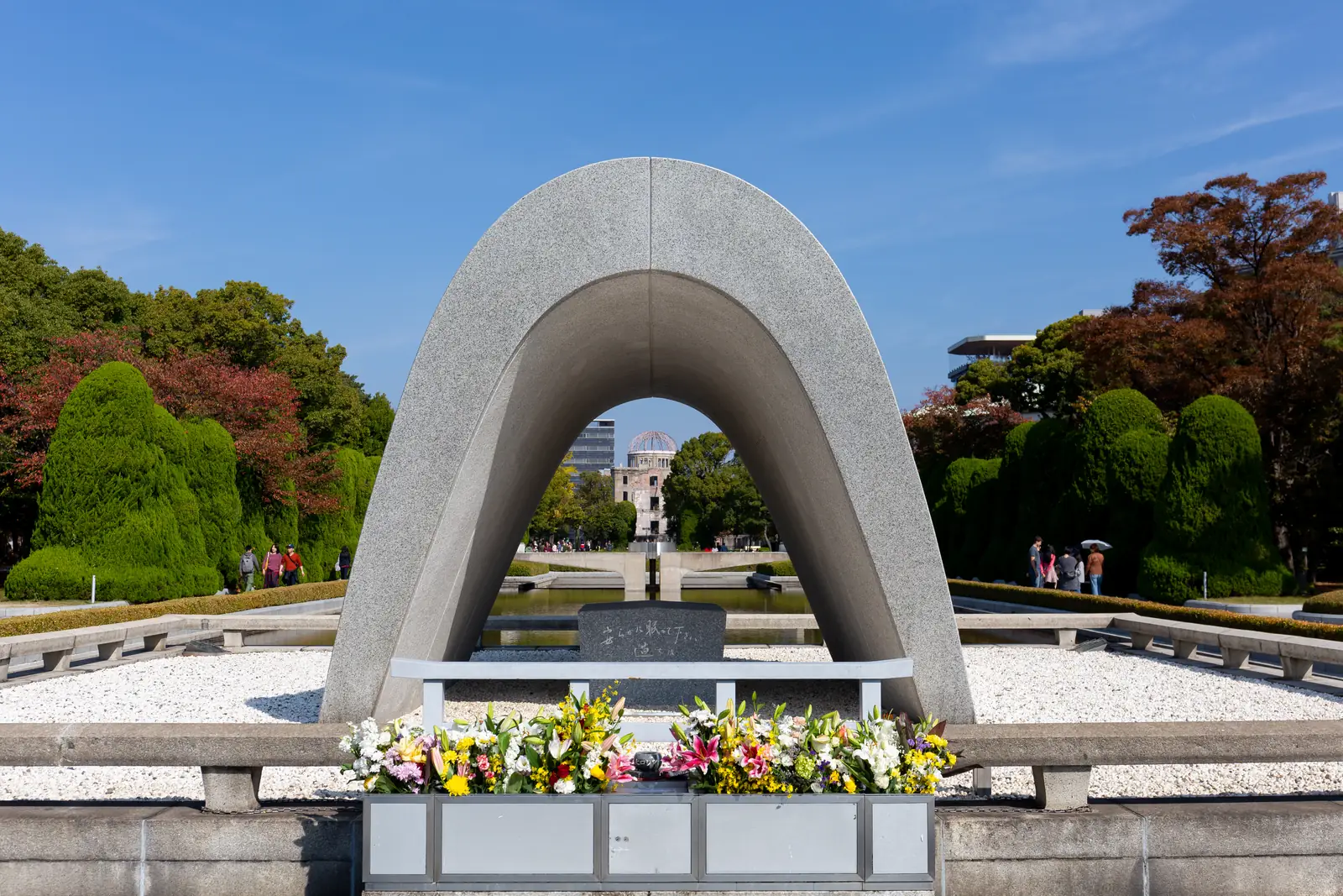 The arch of the Memorial Cenotaph with flowers laid in front and the Atomic Bomb Dome visible through it in the distance