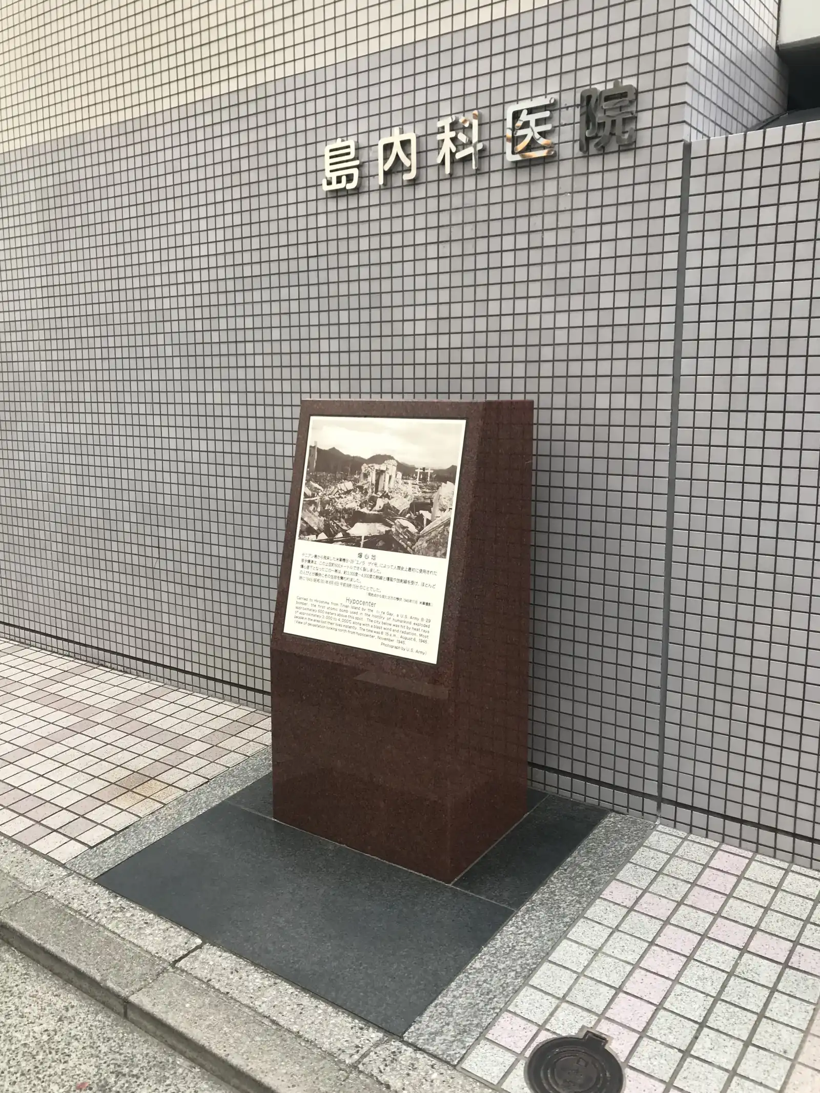 A dark stone marker with an explanatory plaque on the pavement in front of the tiled wall of Shima Hospital, which marks the hypocentre